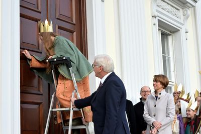 Bundespräsident Frank Walter Steinmeier hält die Leiter für eine Sternsingerin die grade den Segen der Sternsinger an die Pforte des Schlosses Bellevue schreibt. Die restlichen Sternsinger schauen dabei zu. die Frau des Bundespräsidenten freut sich über den Segen. 